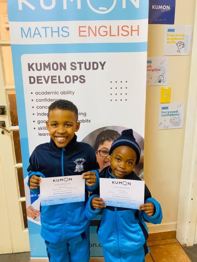 Learners holding Kumon certificates beside a Kumon Mathematics and English display